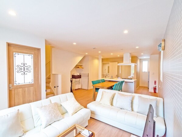 Una sala moderna con un sofá blanco, un comedor con mesa de madera y una cocina al fondo.