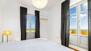 A bedroom with a bed, two windows with curtains, an air conditioning unit, and a view of a yellow building. - Bralos Villa - Paros - Near Naousa, spectacular sea views, 2 bedrooms, privacy (Paros)