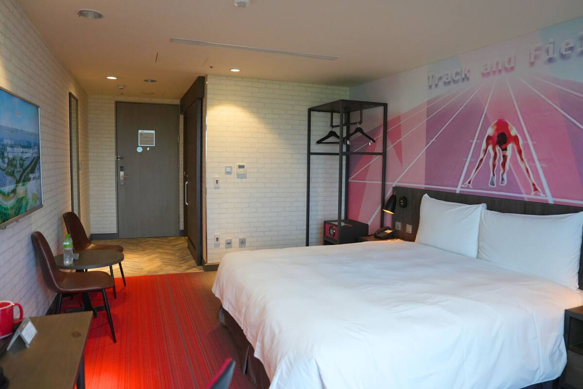 A hotel room with a bed, a desk, a chair, a coat rack, and a wall with a track and field image.