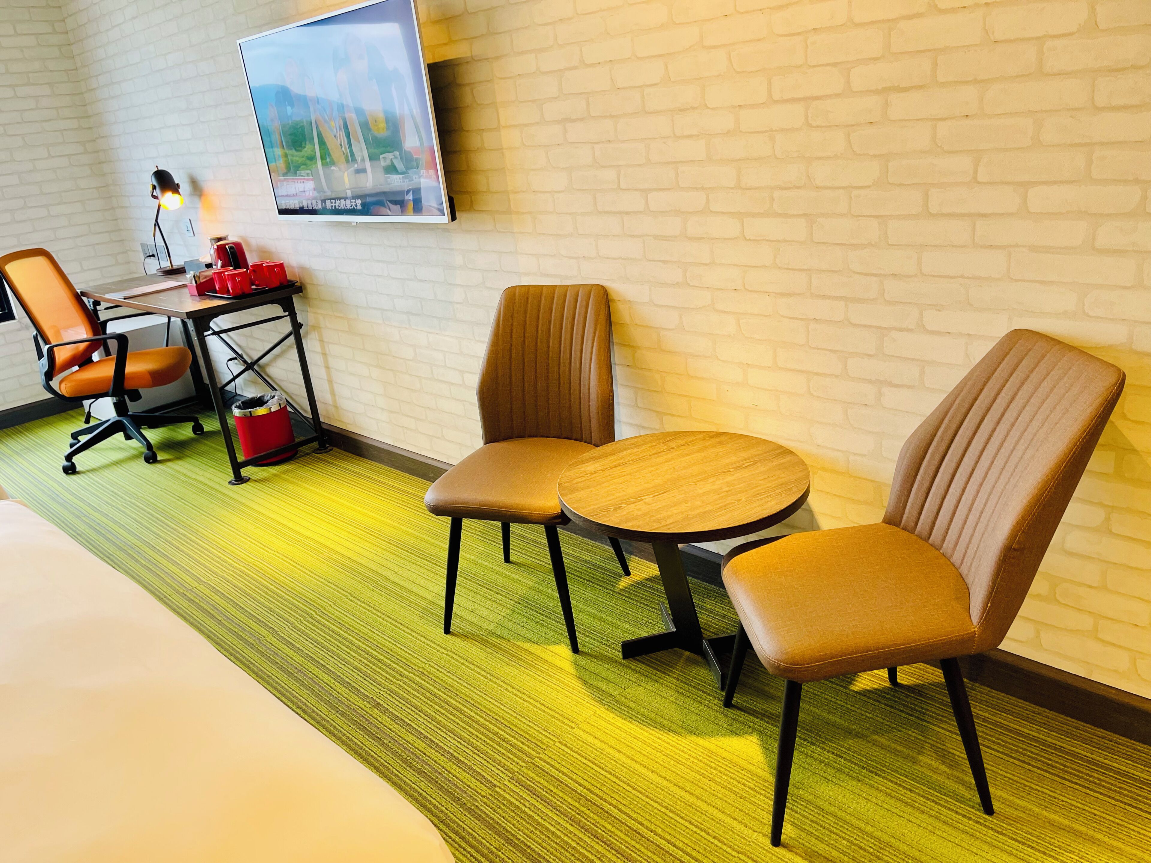 A hotel room with a bed, two chairs, a desk with a lamp, and a TV on the wall.