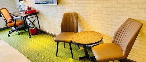 A hotel room with a bed, two chairs, a desk with a lamp, and a TV on the wall.