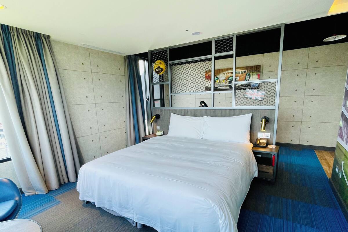 A hotel room with a large bed, a desk, a chair, a nightstand, a lamp, and a window with curtains.