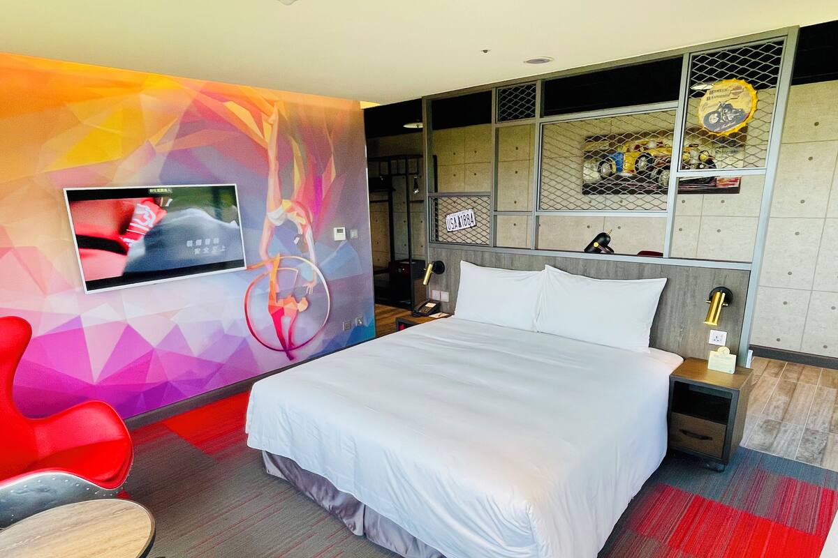 A hotel room with a bed, a TV mounted on the wall, a red chair, and a desk with a lamp.