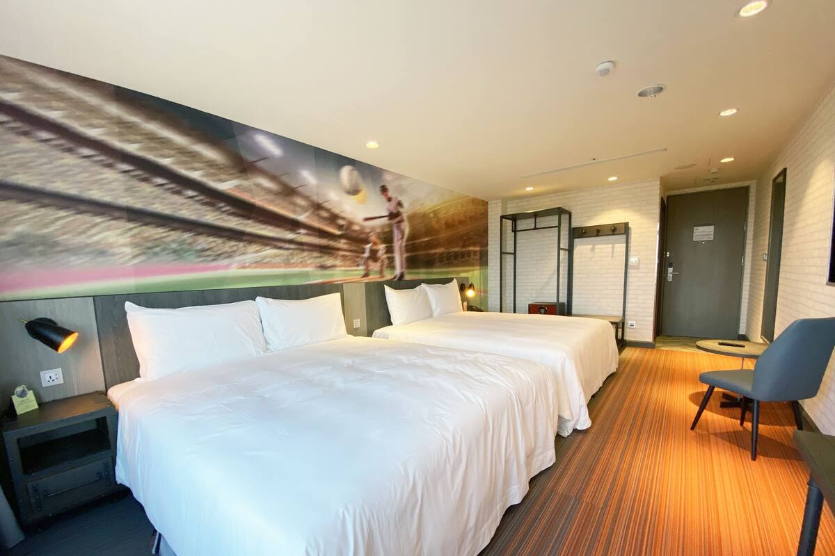 A hotel room with two beds, a desk, a chair, and a mural on the wall.