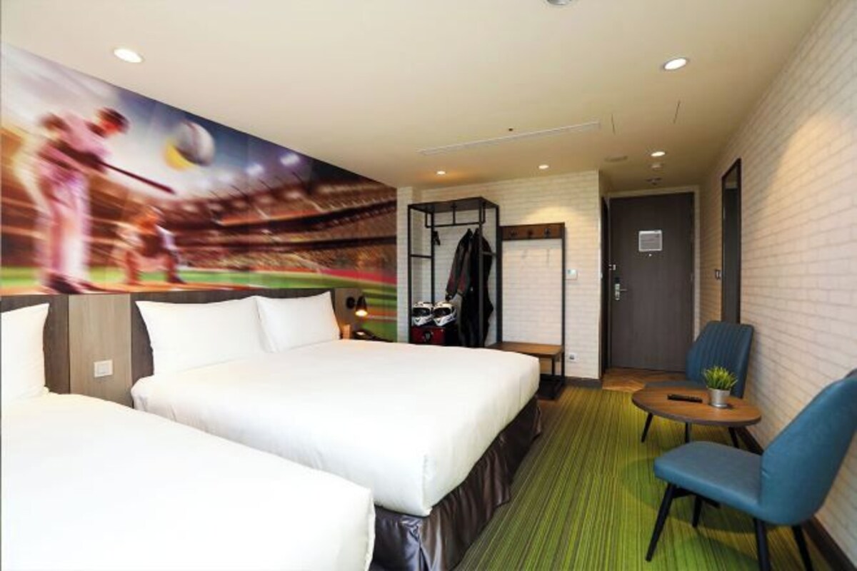 Habitación de hotel con dos camas, una zona de estar con mesa y sillas, un perchero y un mural de un partido de béisbol en la pared.