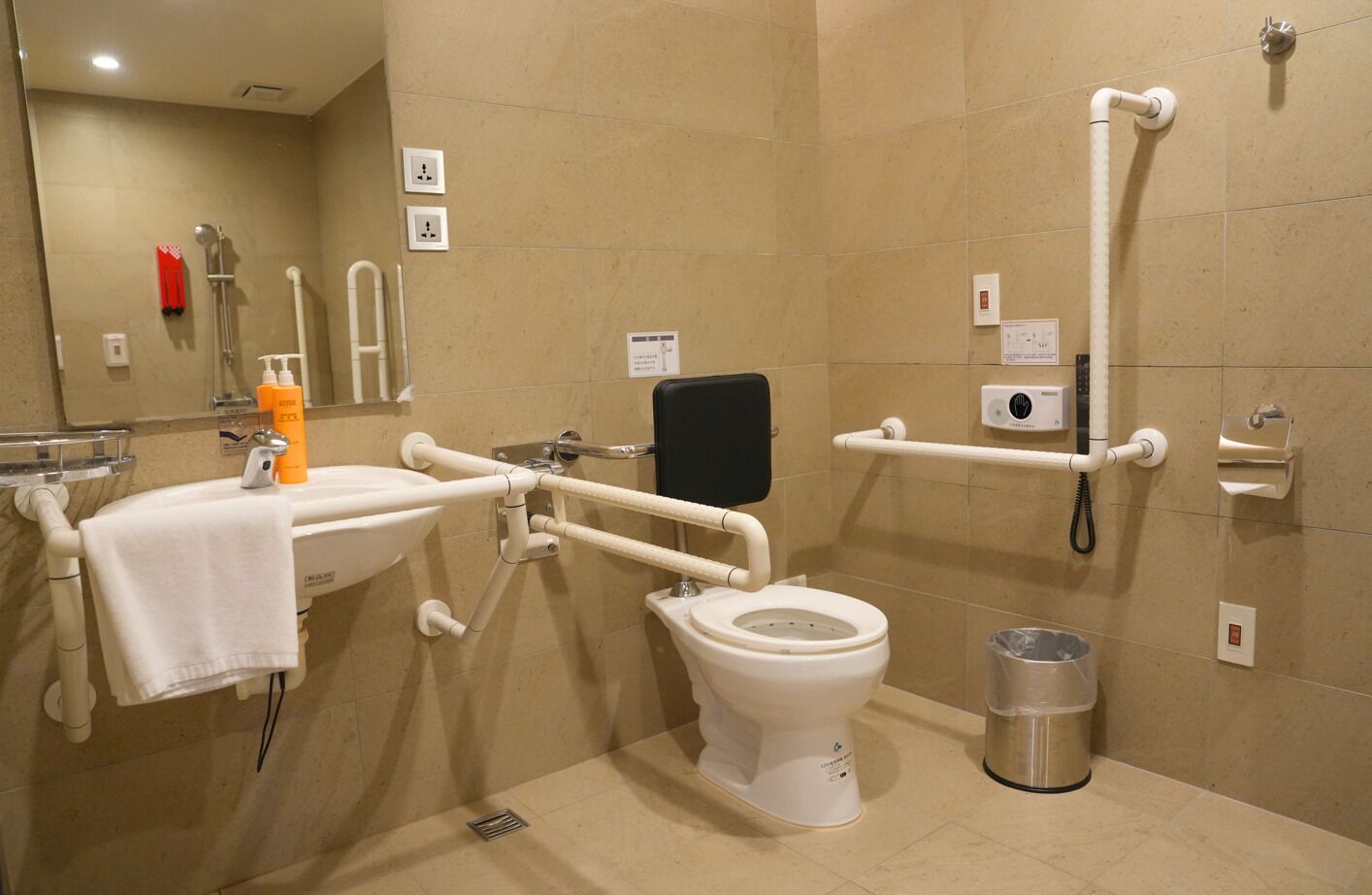 A bathroom with a white toilet, a sink, a handrail, a trash bin, and a shower area.