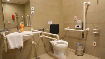 A bathroom with a white toilet, a sink, a handrail, a trash bin, and a shower area.
