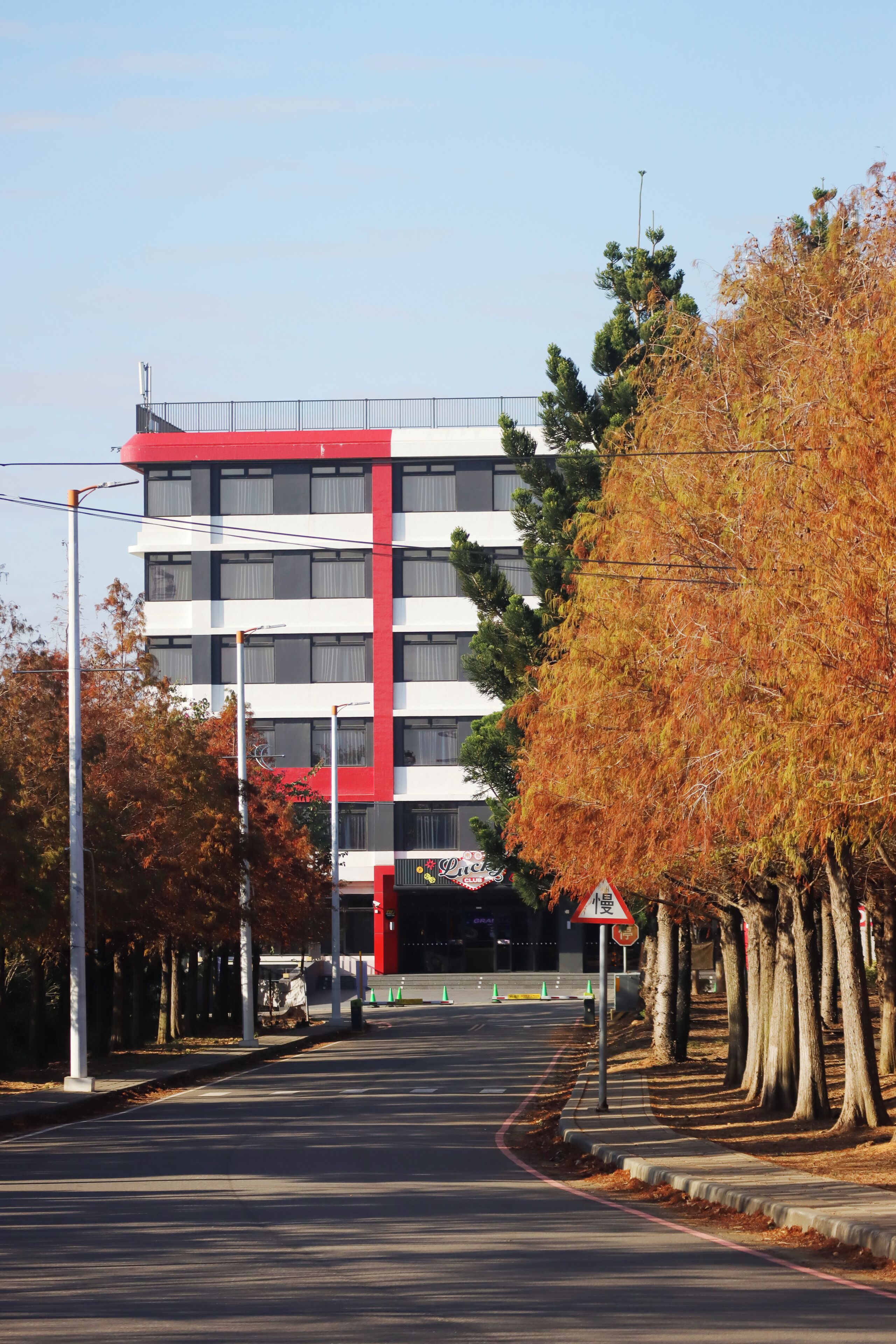 A multi-story building with a red and white facade, a clear road in front, and trees with autumn foliage.