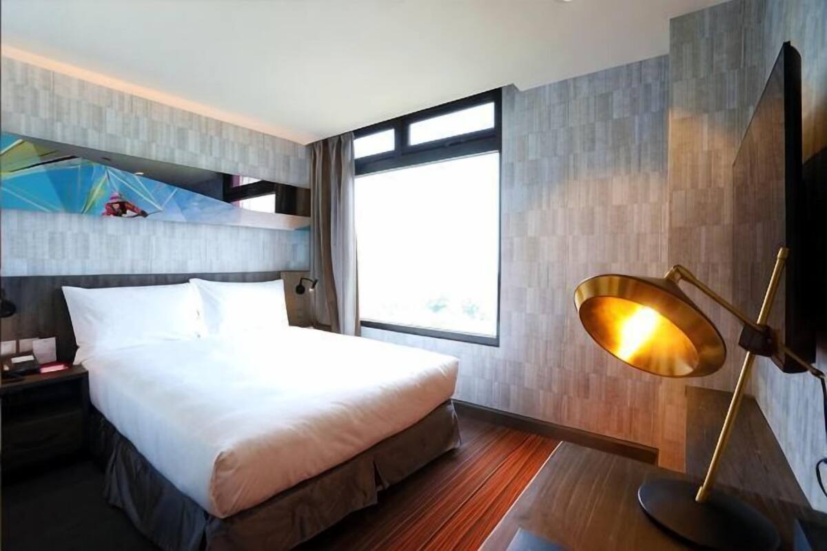 A hotel room with a bed, a desk, a lamp, and a large window.