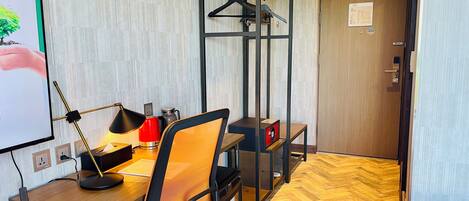 A hotel room with a wooden desk, an orange office chair, a black hanging rack, and a bed with a white cover.