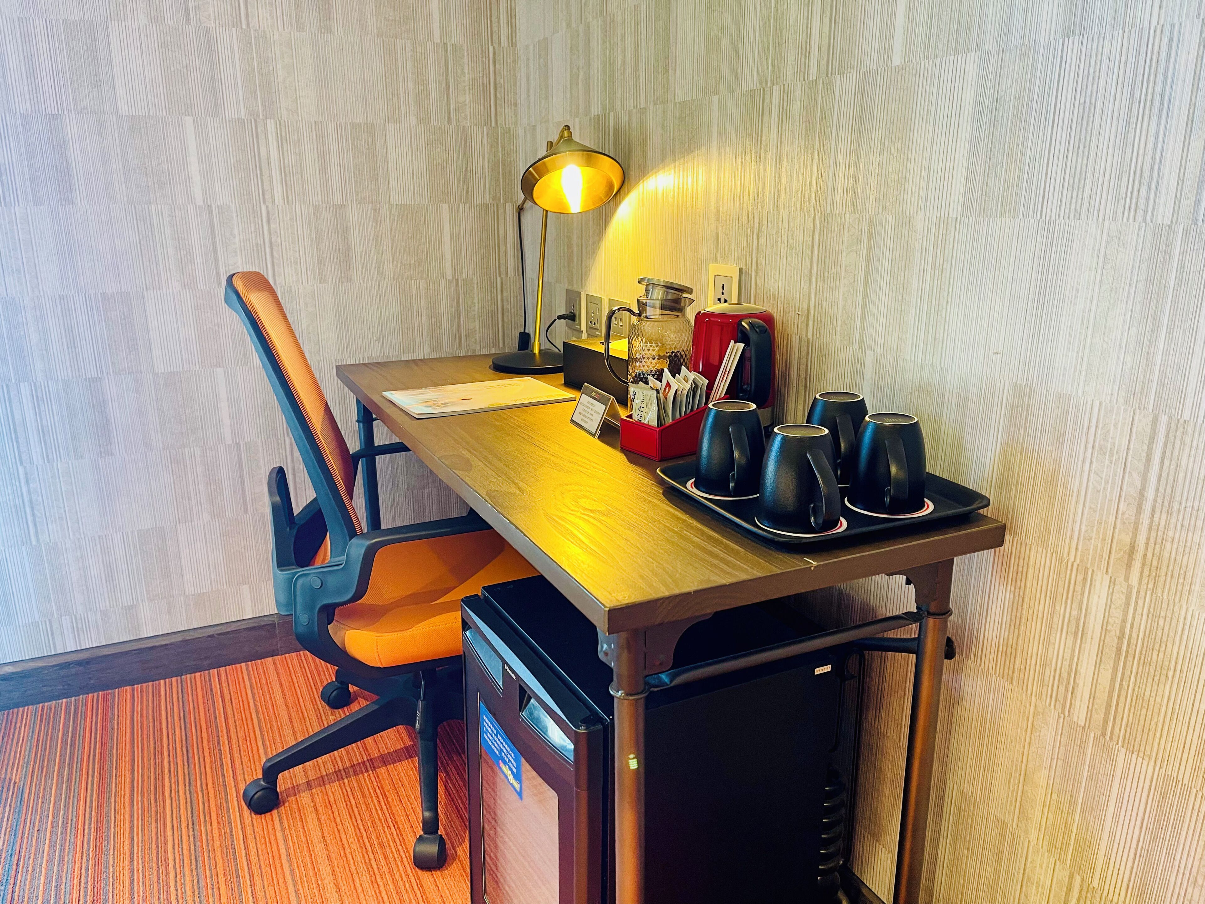 Habitación de hotel con un escritorio de madera, una silla, una lámpara, una cafetera y un refrigerador.