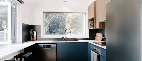 A modern kitchen with a large window, dark blue cabinets, a stainless steel dishwasher, and a black refrigerator.