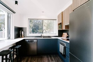 Une cuisine moderne dotée d’une grande fenêtre, de meubles bleu foncé, d’un lave-vaisselle en acier inoxydable et d’un réfrigérateur noir.