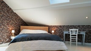 A bedroom with a sloped ceiling, a wooden headboard, a bed with a white and dark blue quilt, a desk with a chair, and a brick wall. - Gîte la maisonnette in the heart of the Loire (L'hôpital le grand)