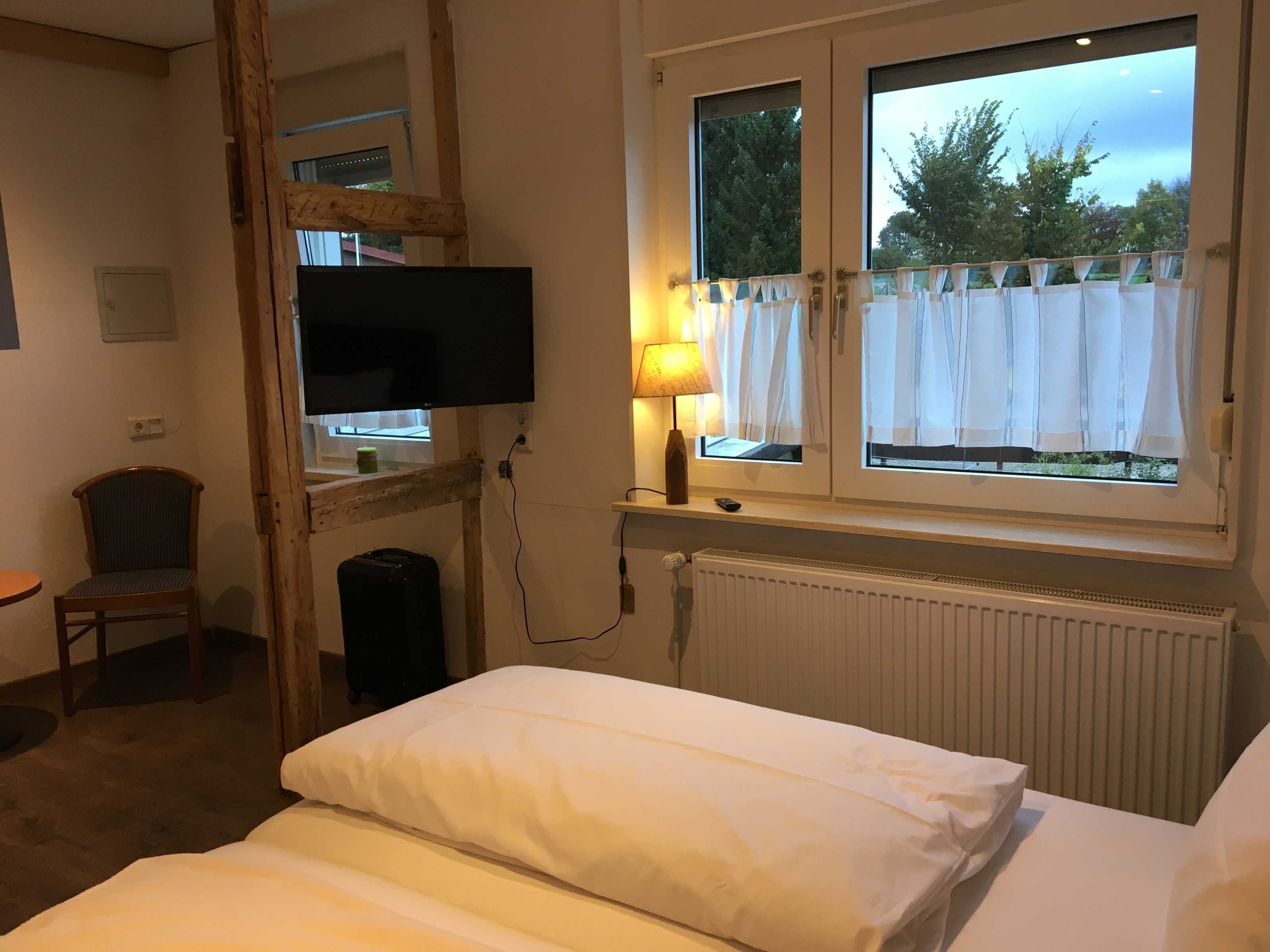 A hotel room with a bed, a chair, a TV mounted on a wooden beam, a window with curtains, and a radiator.