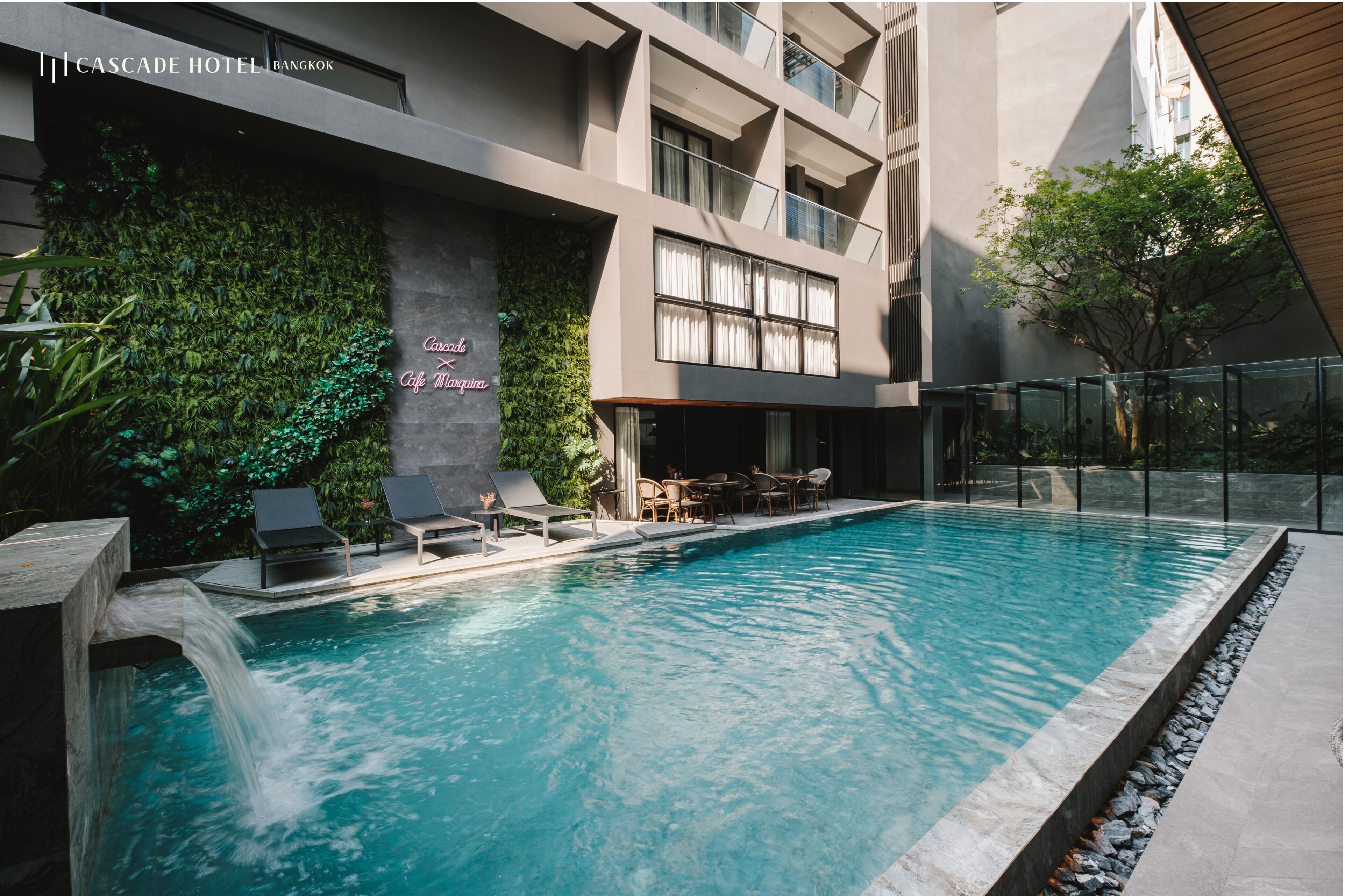 A modern hotel pool area with a waterfall feature, lounge chairs, and a seating area with a view of the building's interior.