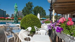 An outdoor seating area with white plastic chairs and tables, a pool in the background, and potted flowers along the railing. - Aparthotel Poseidon - All inclusive (Sunny Beach)