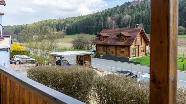 Una casa de madera con garaje y porche, rodeada de un paisaje rural con carretera y coches aparcados.
