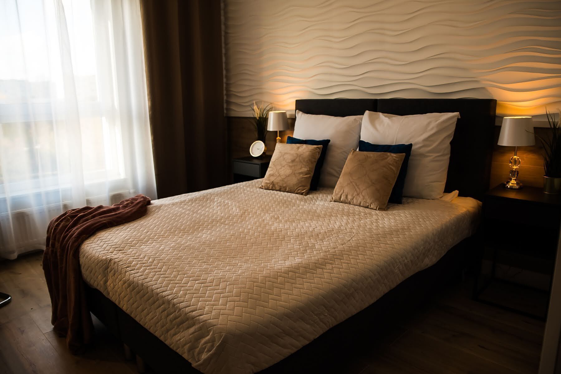 Cama bem arrumada com cobertura acolchoada, duas luminárias de cabeceira e uma parede com textura.