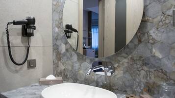 A bathroom with a marble countertop, a white sink, a round mirror, and a hairdryer mounted on the wall.