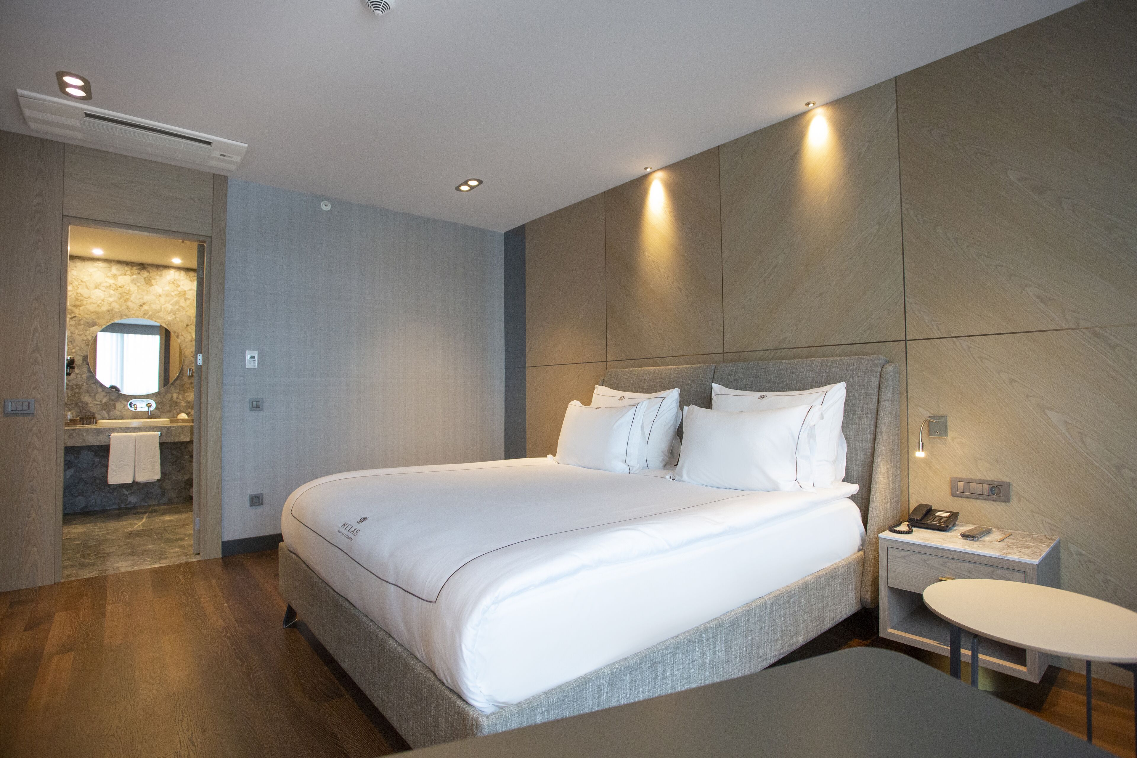 Una habitación de hotel moderna con una cama grande, una mesita de noche con teléfono y lámpara, y una pequeña mesa redonda.