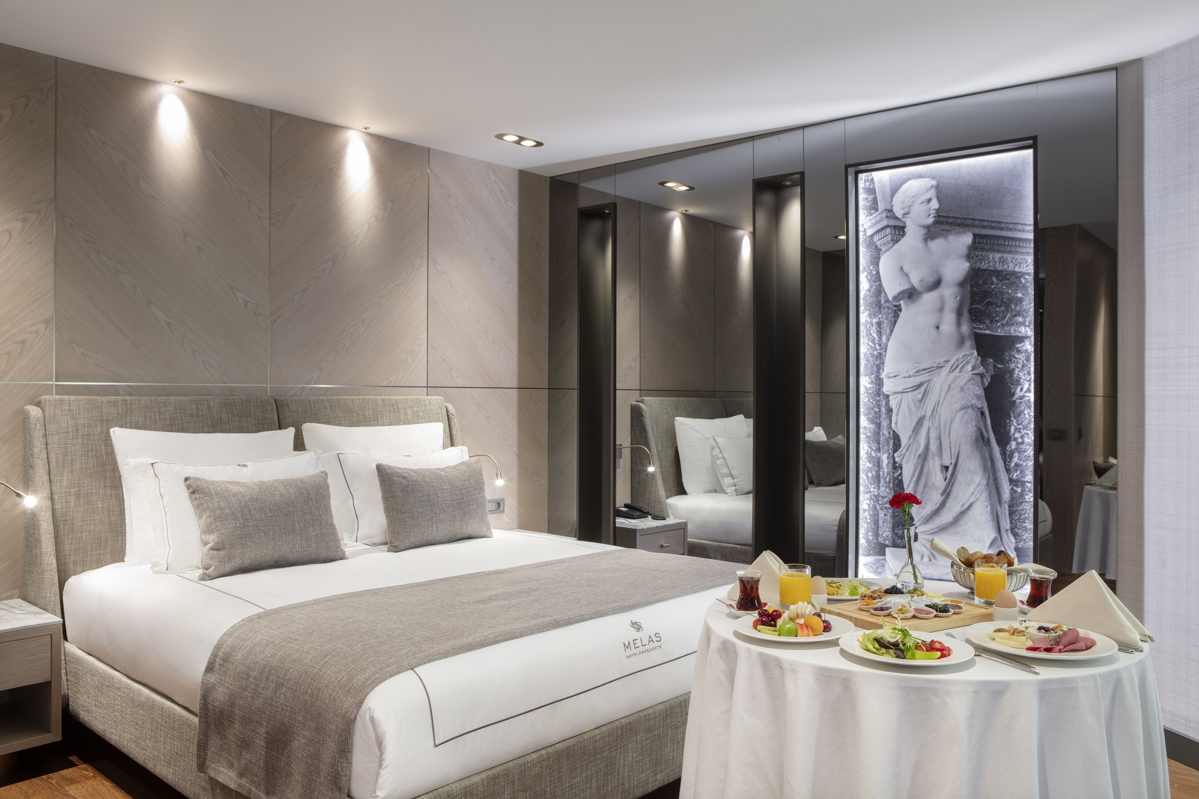 A modern hotel room with a large bed, a dining table with breakfast, and a large artwork on the wall.
