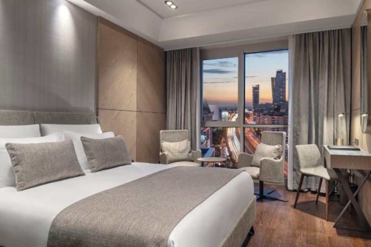 A hotel room with a large bed, two armchairs, a small table, and a view of a cityscape at sunset.