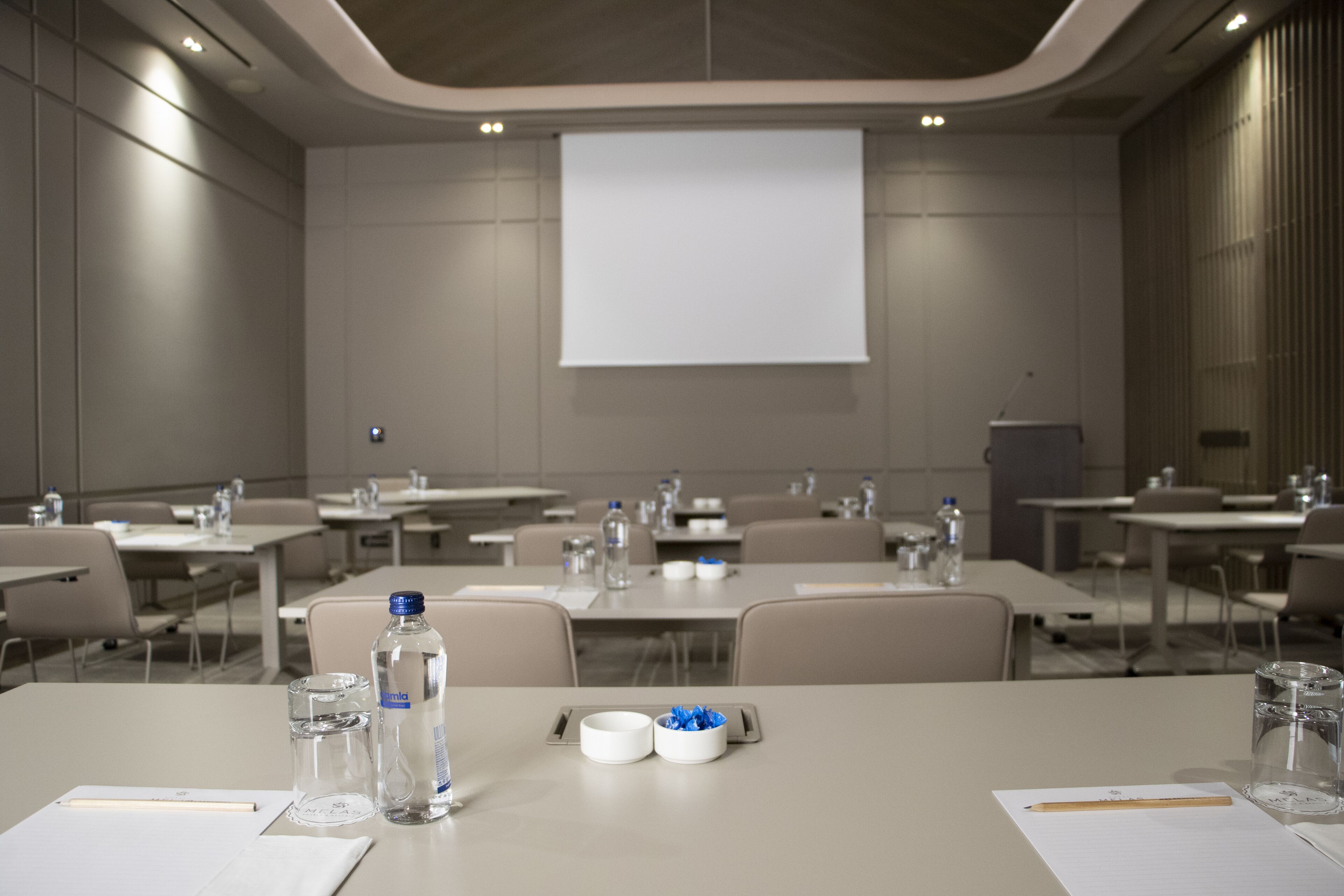 Un salón de conferencias con mesas, sillas, botellas de agua y una pantalla de proyector.