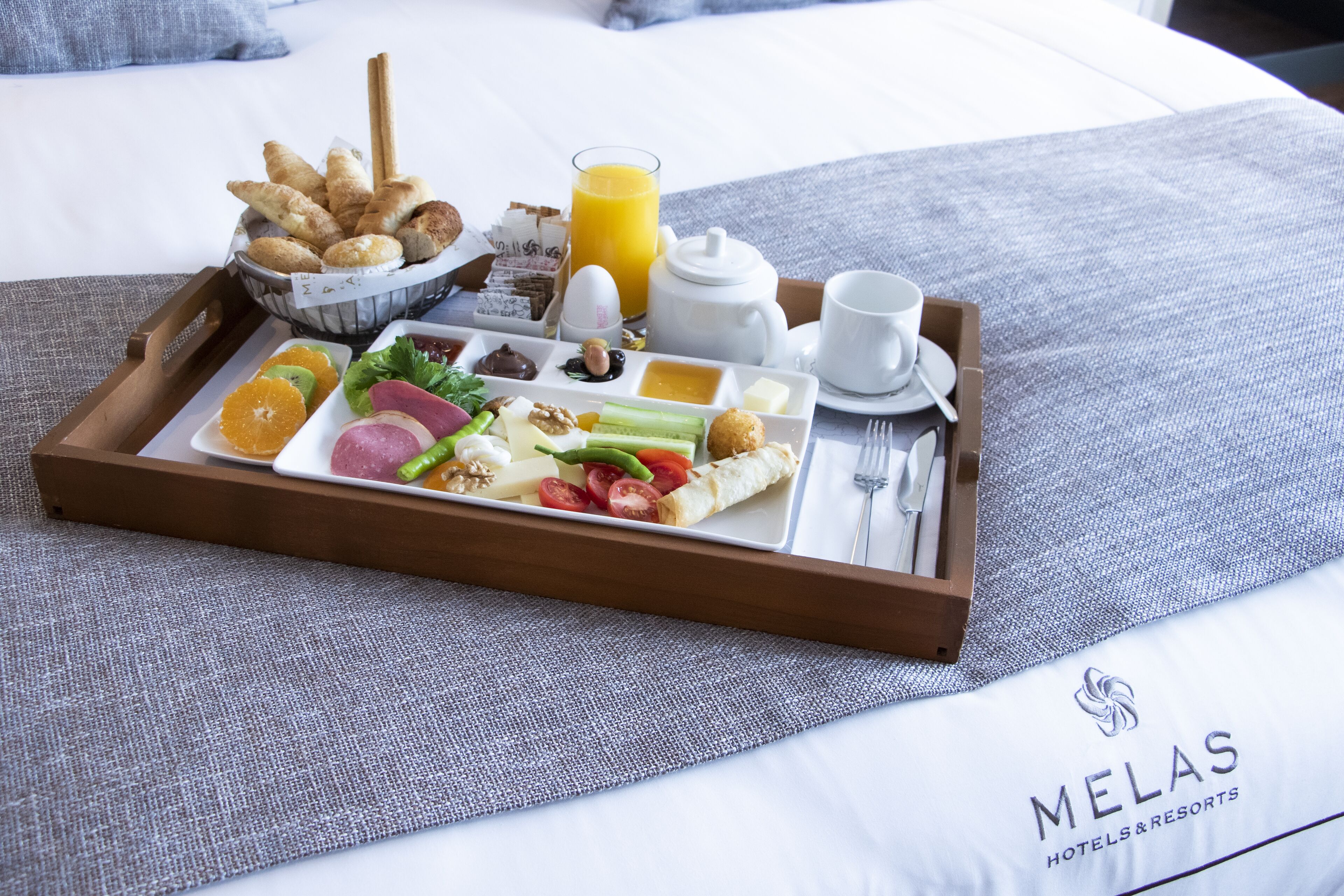 A breakfast tray with assorted foods and a glass of orange juice on a bed with a gray pillow and a white sheet.