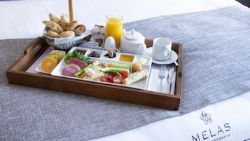 A breakfast tray with assorted foods and a glass of orange juice on a bed with a gray pillow and a white sheet.