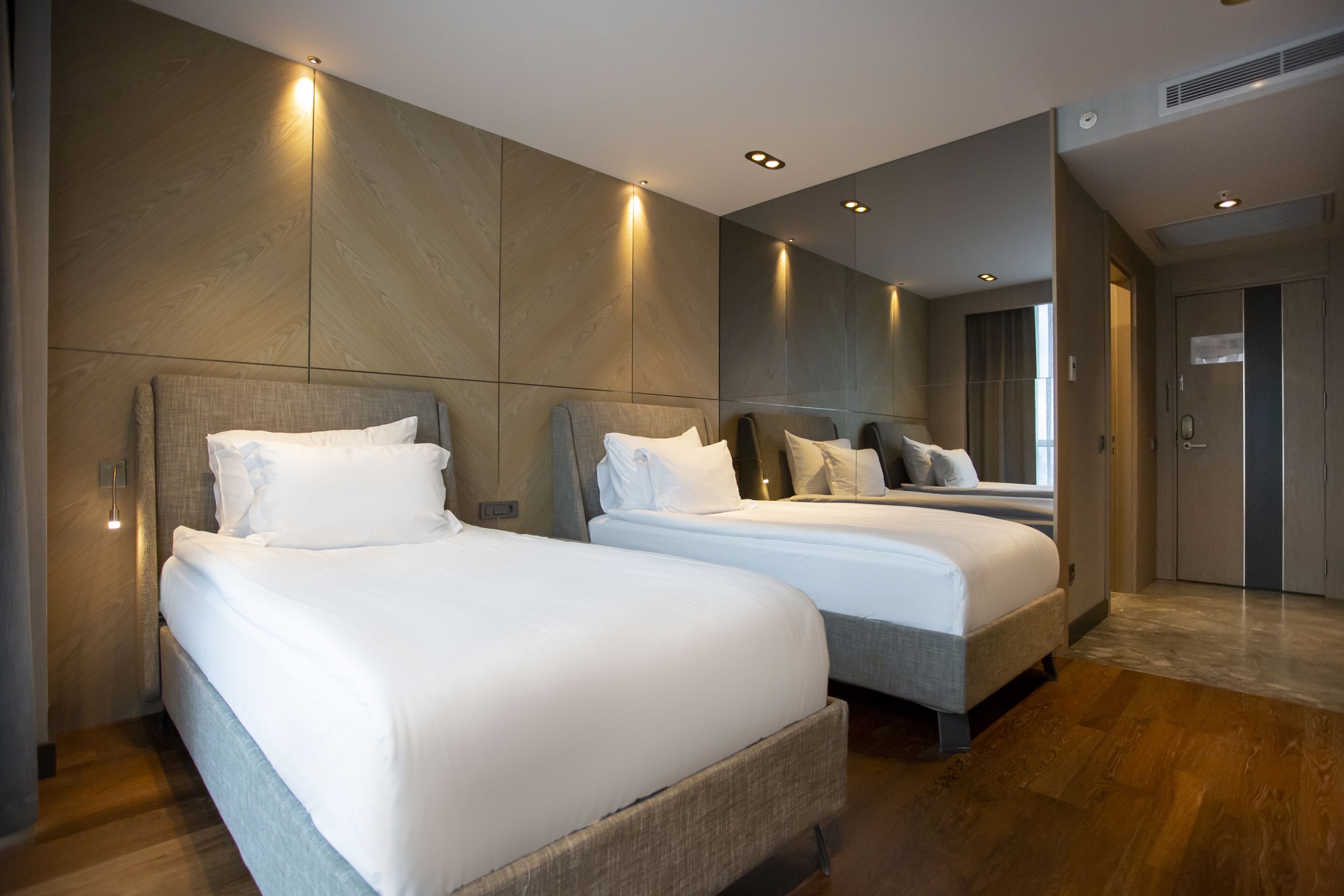 A modern hotel room with two beds, wooden flooring, and a large mirror on the wall.