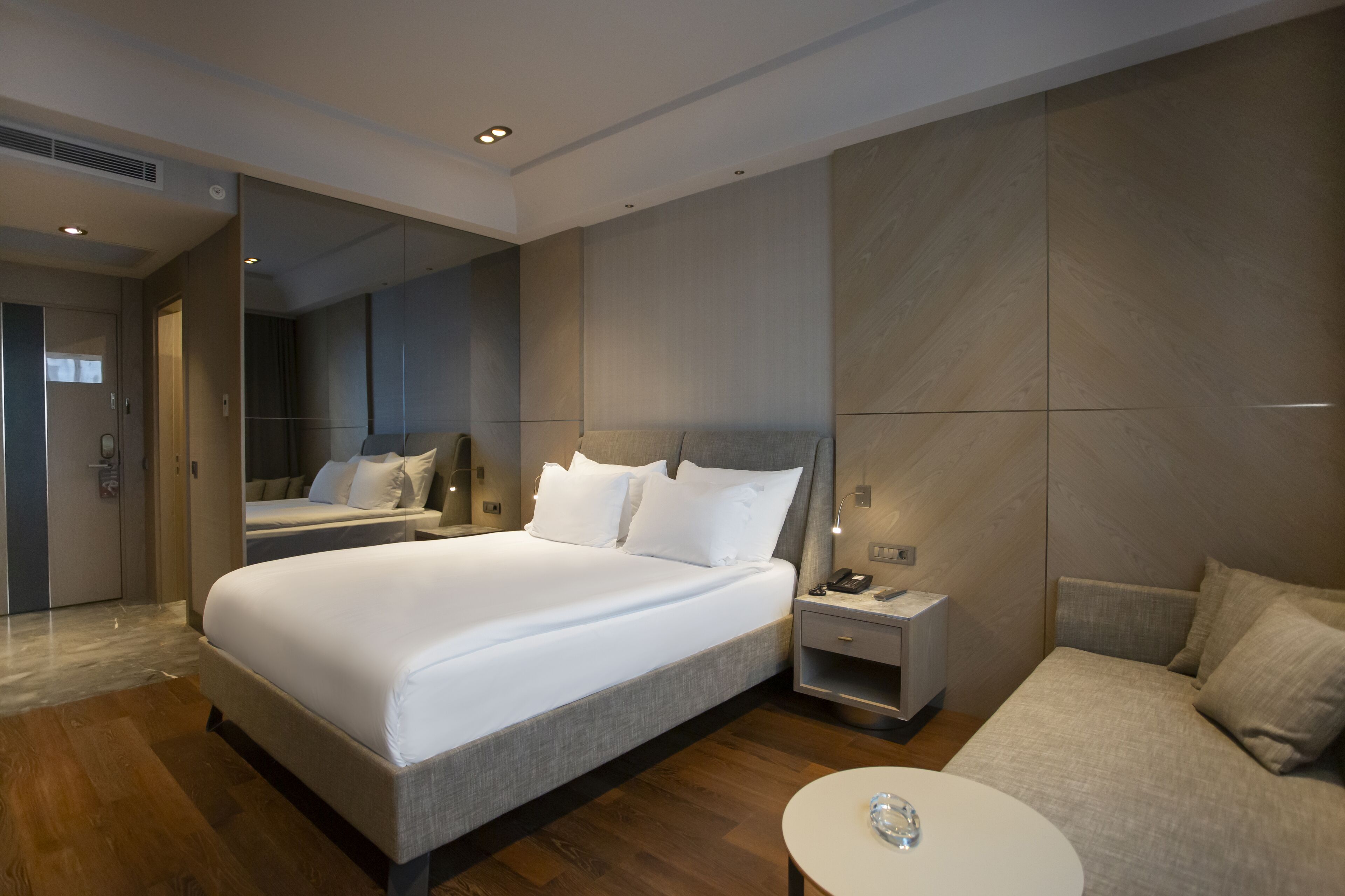 A modern hotel room with a large bed, a bedside table with a phone, a small bedside table with a lamp, and a round table with a decorative item.