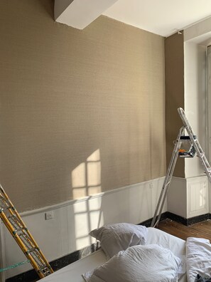 A room with a ladder leaning against a wall, a window with sunlight casting shadows, and a bed with pillows.