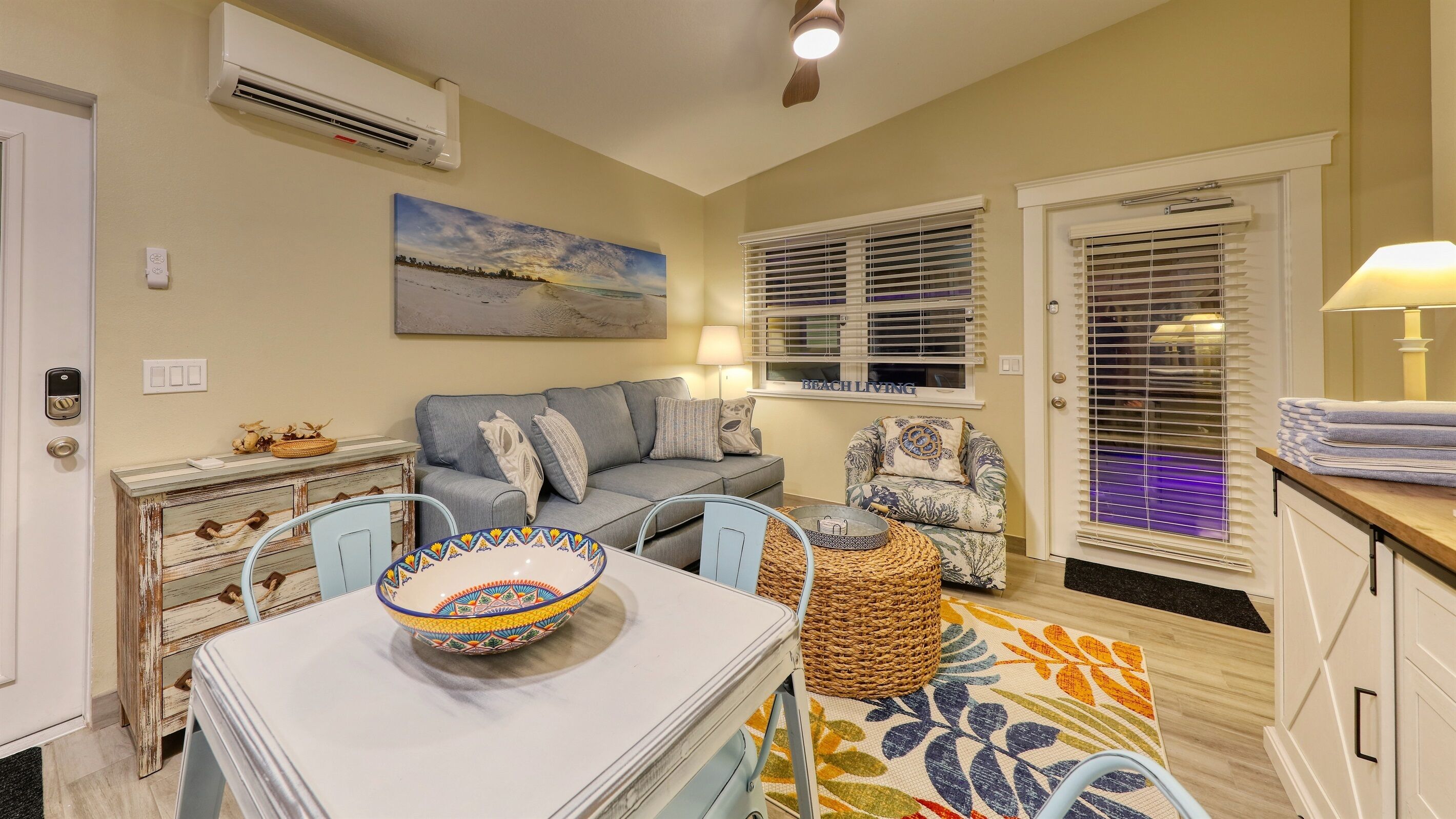 A compact living room with a sofa, a dining table with a bowl, a chair, a small side table, and a window with blinds.