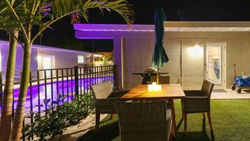 Une terrasse avec une table et des chaises, une piscine éclairée de lumières violettes et un espace abrité avec un barbecue.
