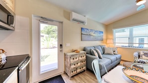 Un espacio habitable compacto con kitchenette, un sofá gris, un cuadro y una ventana con persianas.