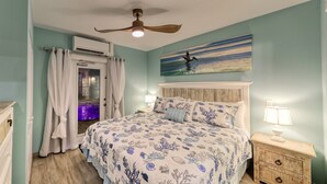 A bedroom with a bed, a ceiling fan, an air conditioning unit, a nightstand with a lamp, and a painting on the wall.