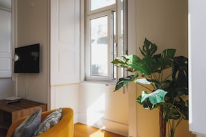 Une pièce avec un plancher en bois, un canapé jaune, une grande plante verte et une fenêtre donnant sur des immeubles.