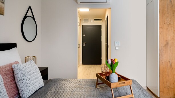 A modern bedroom with a bed, a bedside table, a wooden bench with a vase of flowers, and a door leading to another room. - EASY RENT Apartaments - SMART 318 (Lublin)