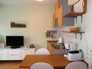 A compact kitchen with a wooden table, white chairs, a television, and basic appliances.