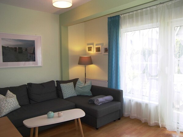 Een woonkamer met een grijze bank, een klein wit tafeltje en een lamp. Er hangen afbeeldingen in lijsten aan de muur en een raam met gordijnen.