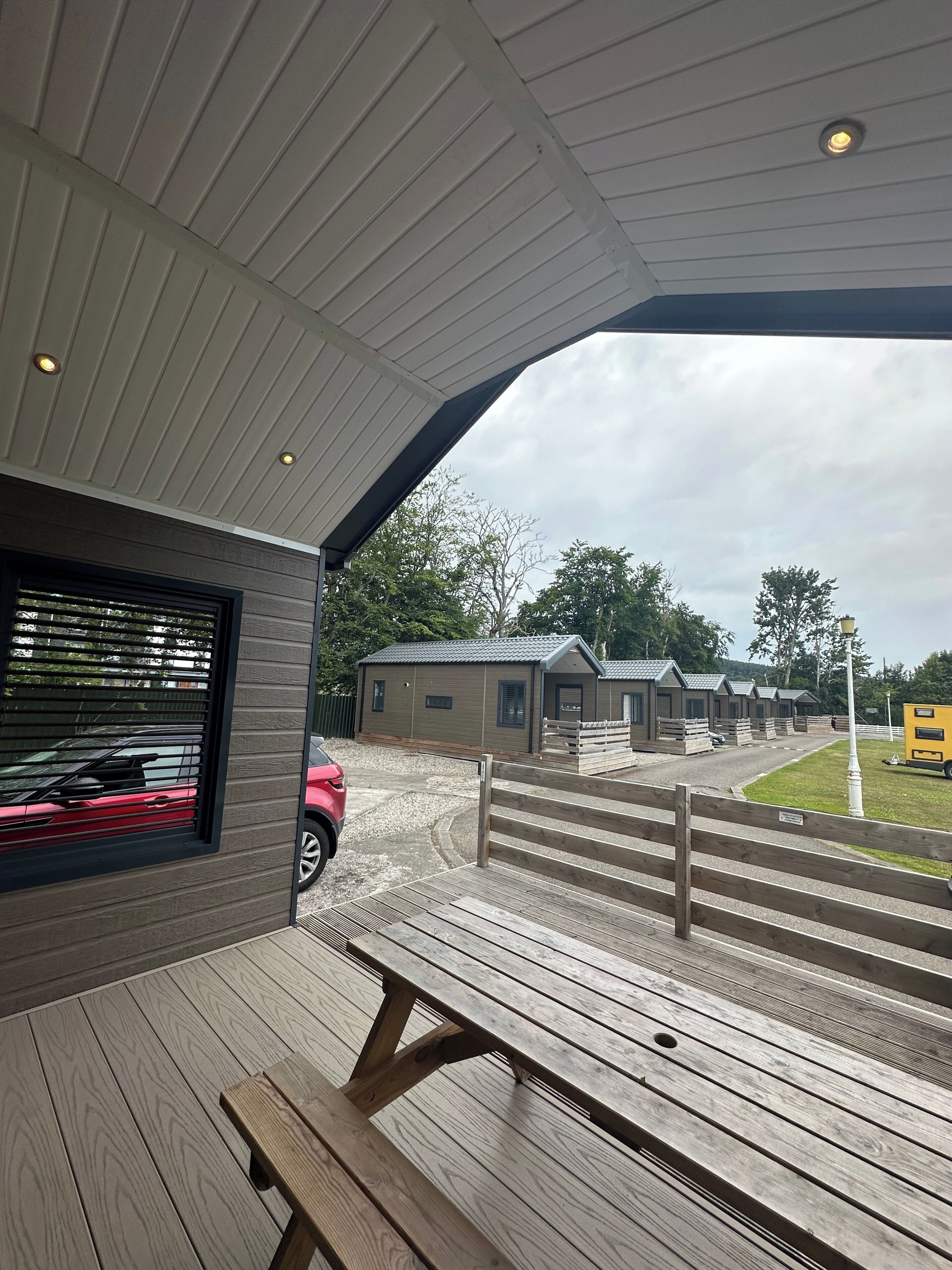 Un espace extérieur abrité, doté de bancs en bois, d’où l’on peut admirer les écuries et, au loin, une voiture rouge.