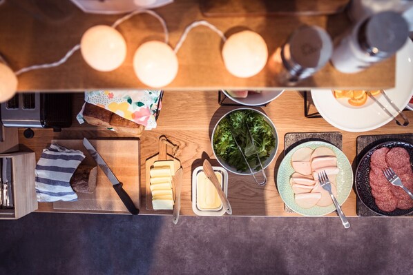 A top-down view of a kitchen counter with various food items and utensils, including a loaf of bread, a cutting board, a cheese slicer, a bowl of salad, and plates with sliced meats. - Hostel Café Koti (Rovaniemi)