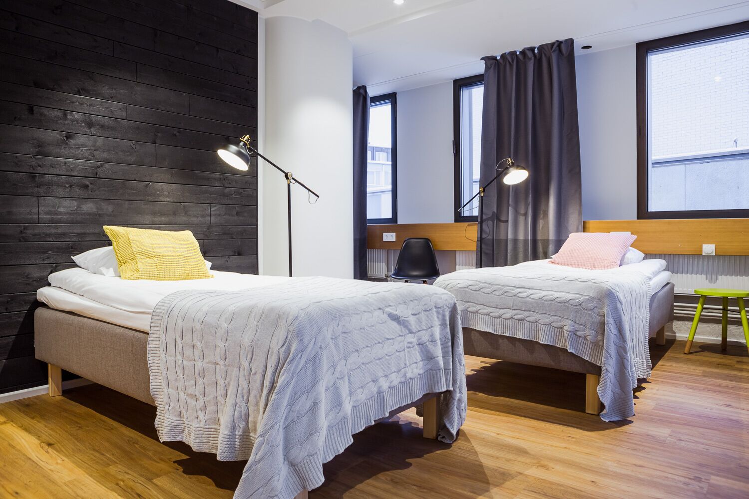 Habitación de hotel moderna con dos camas, suelo de madera, ventanas amplias y una pared negra.