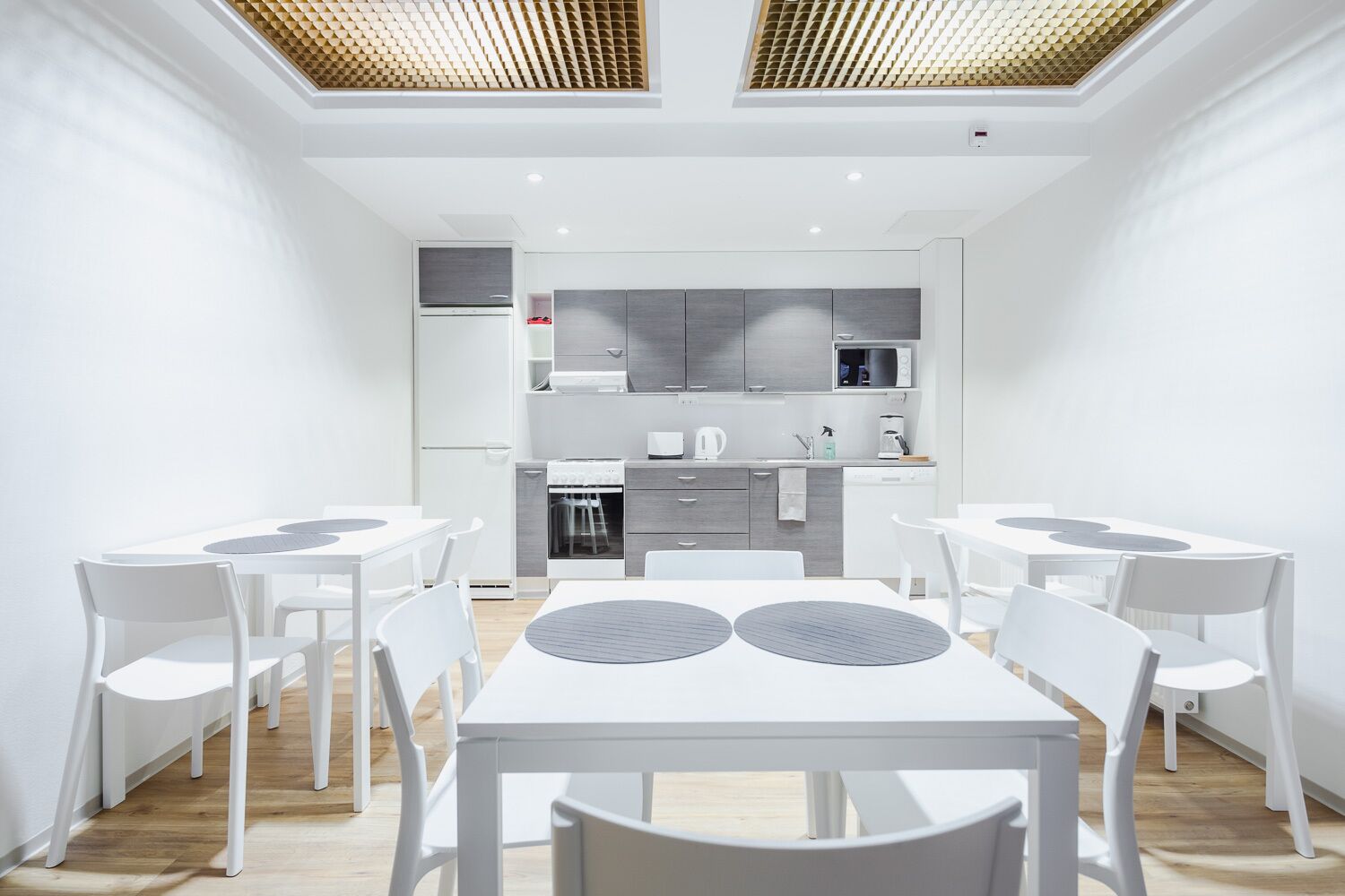 Una cocina moderna con armarios blancos, un microondas y una estufa. Hay dos mesas de comedor blancas con sillas, y un techo con detalles de madera.
