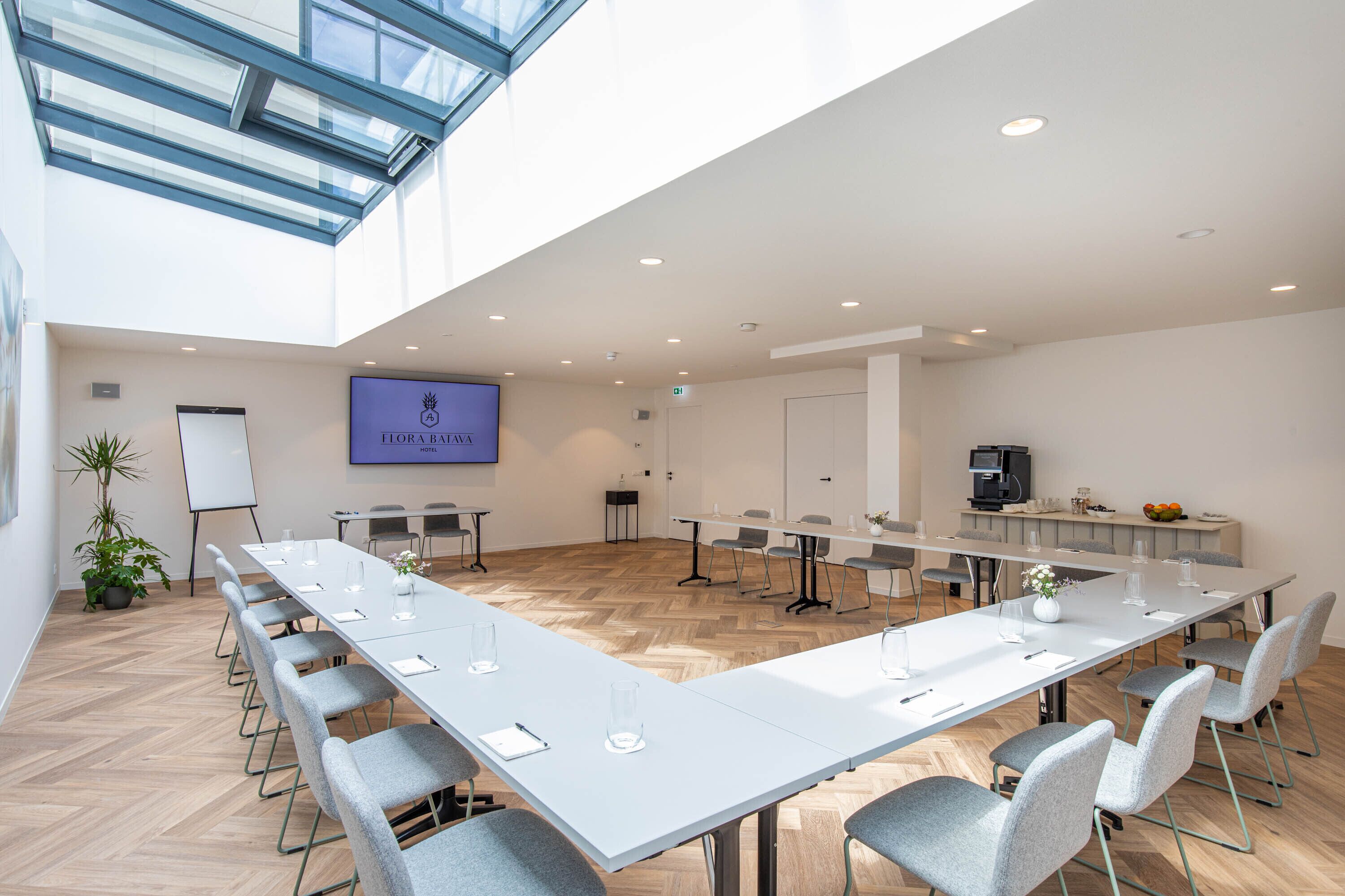 A conference room with a long table, chairs, a whiteboard, and a screen displaying 'FLORA BAVARA'.