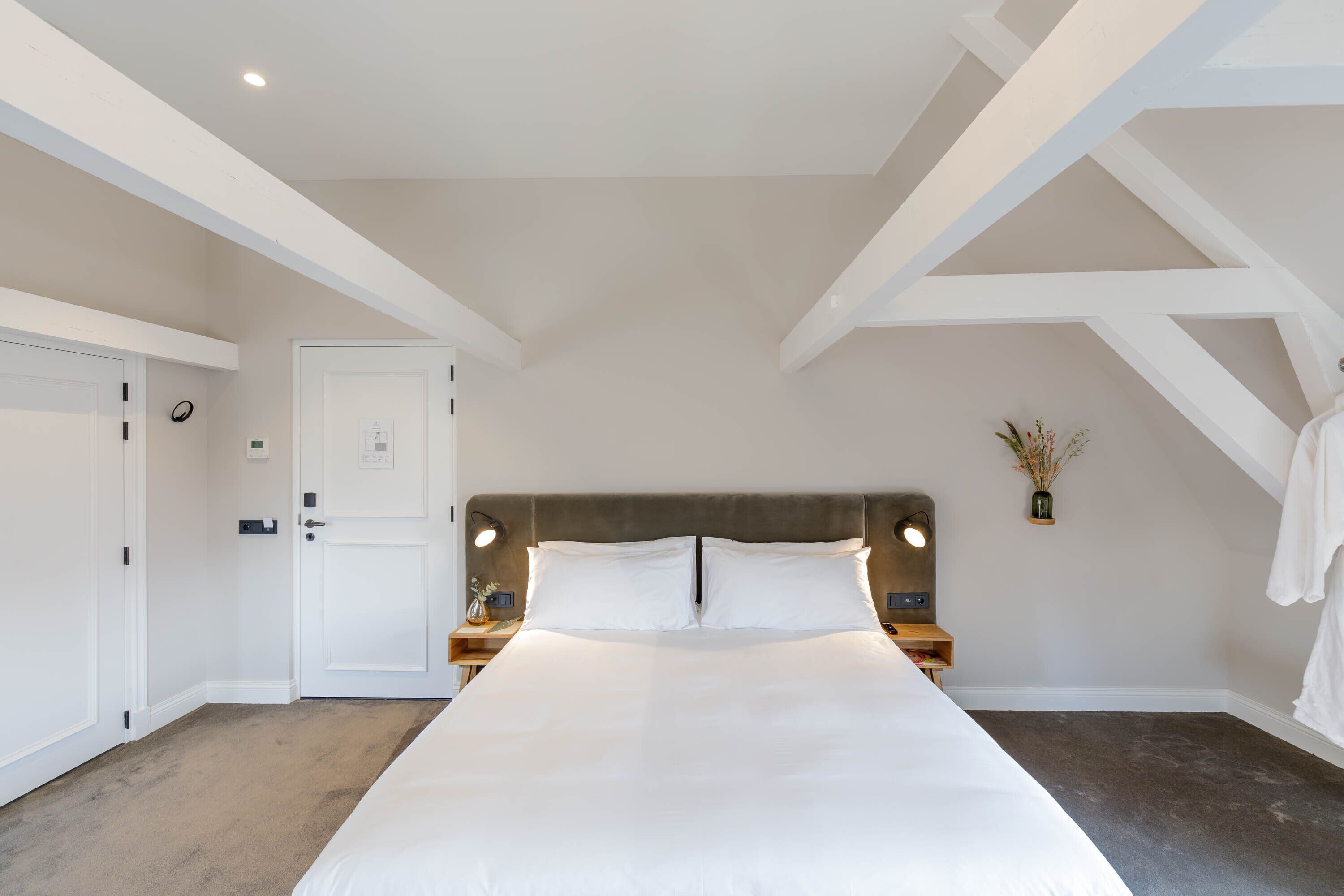 A bedroom with a large bed, white walls, and exposed wooden beams. There are two bedside tables with lamps, a door, and a small vase with dried flowers on the wall.