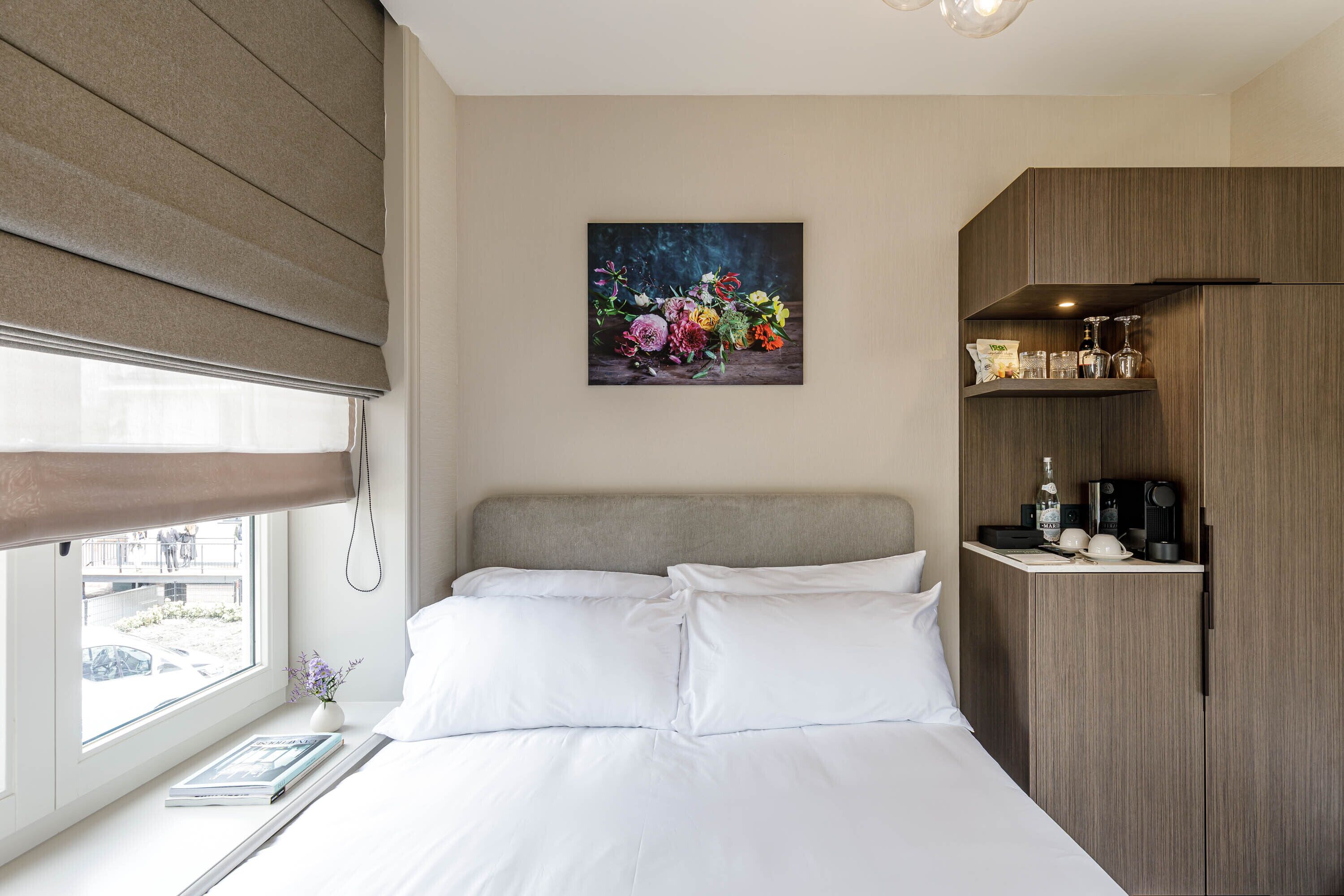 Una habitación de hotel con una cama, un cuadro de flores en la pared, una cafetera y un minibar.