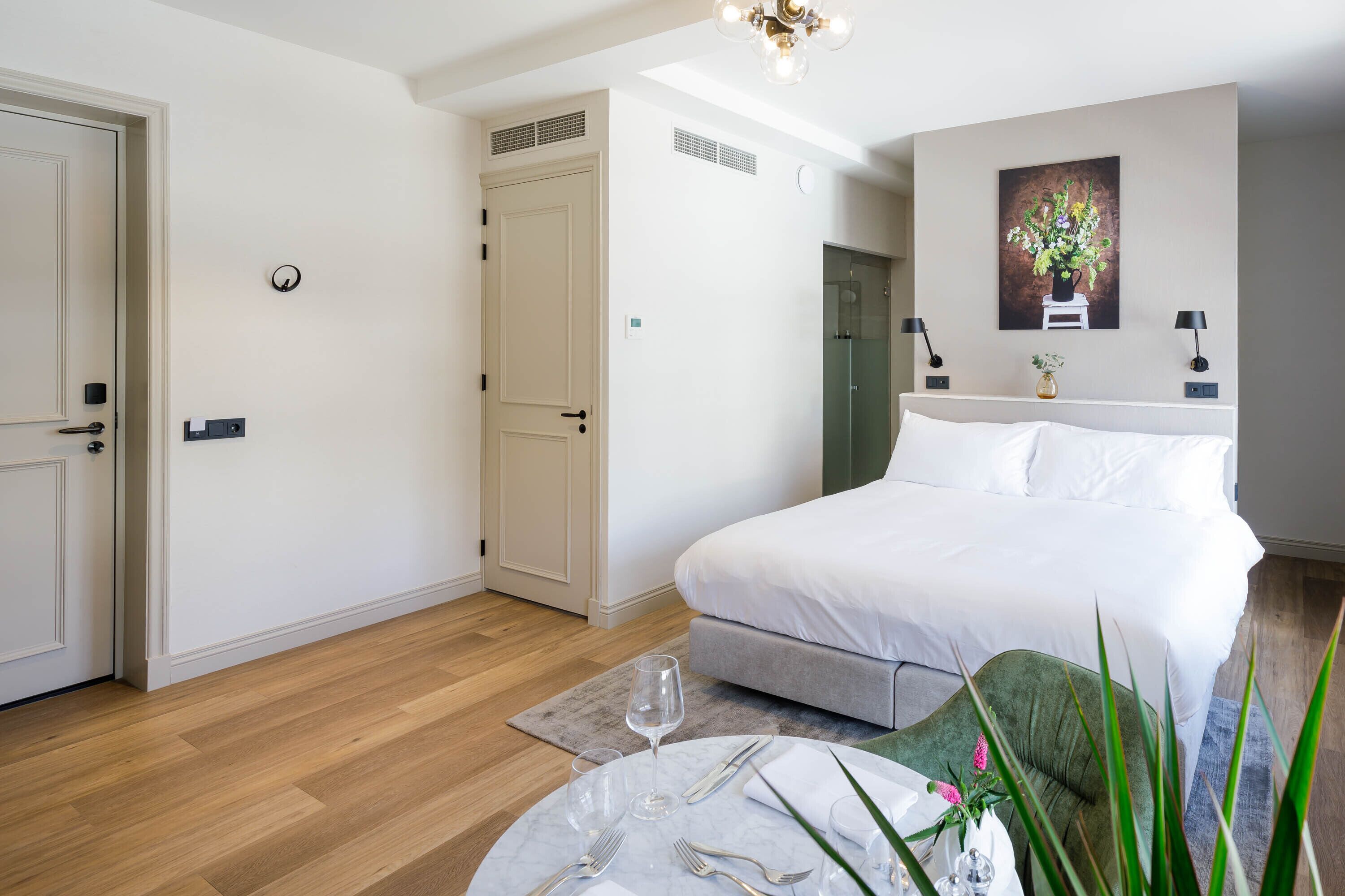 A bedroom with a bed, bedside tables, a round dining table with cutlery, a vase with flowers, and a painting on the wall.