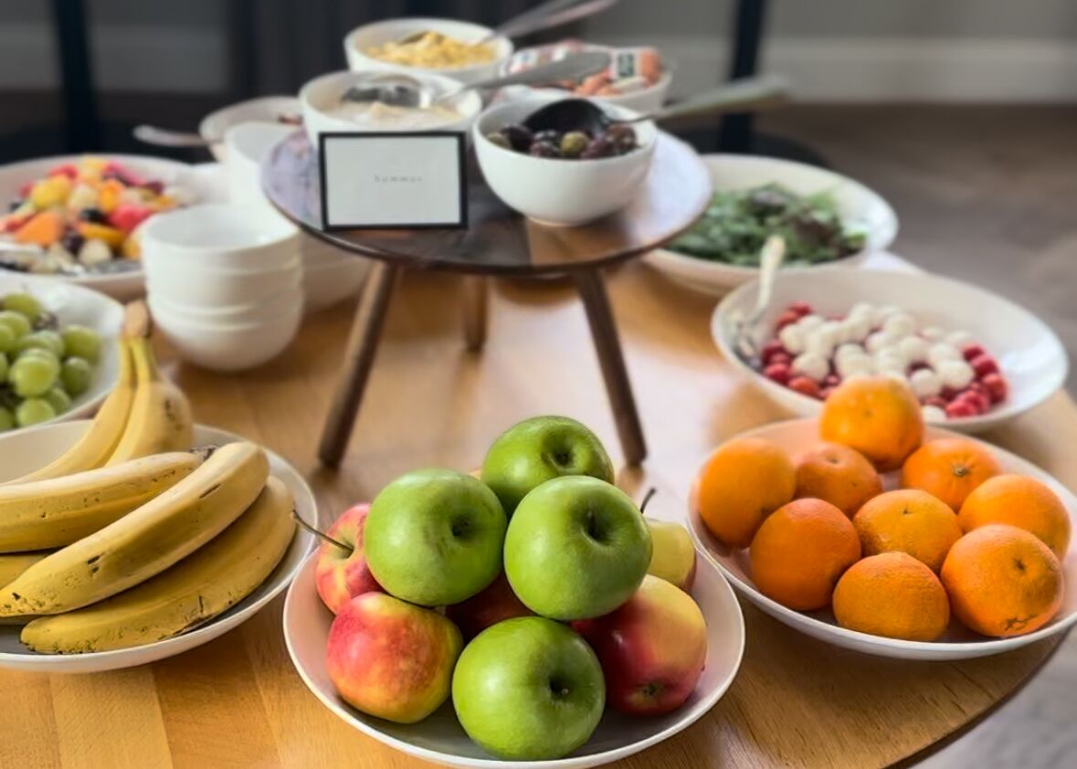 Una mesa con frutas variadas y recipientes de comida, incluyendo plátanos, manzanas, naranjas y bayas.